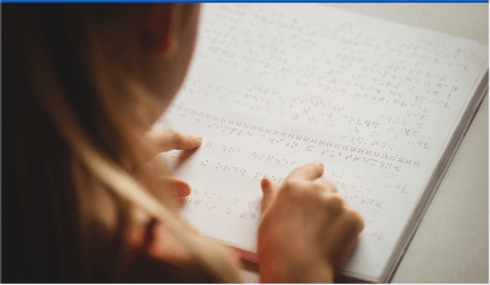 Photo over a young girl's shoulder as she sits at a desk and reads from an open braille book.