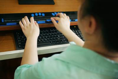 Photo looking over a woman's shoulder as she sits at a computer and uses a braille assistive device