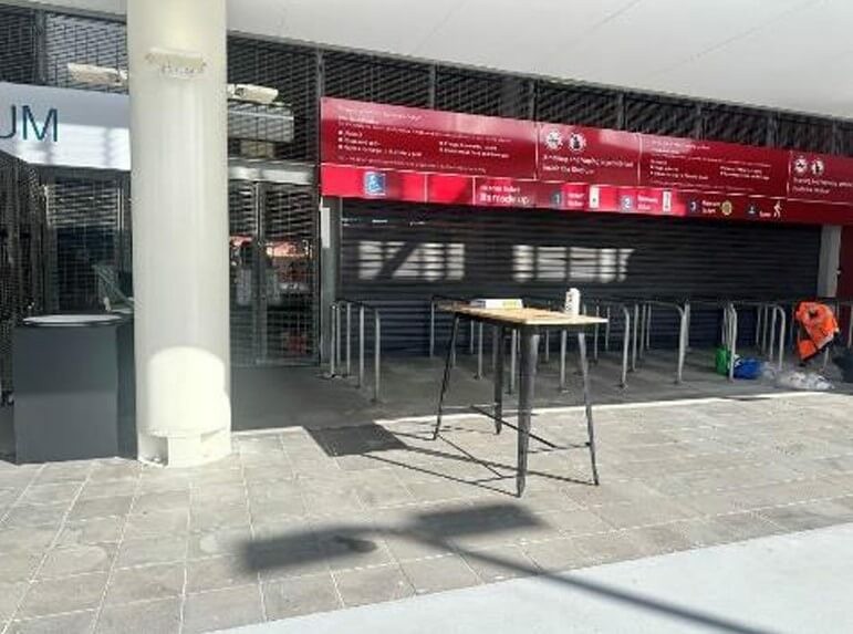 Photograph of Entry area. Lack of Braille or tactile signage. No colour contrast between pole and surrounds. Table is blocking walkway.